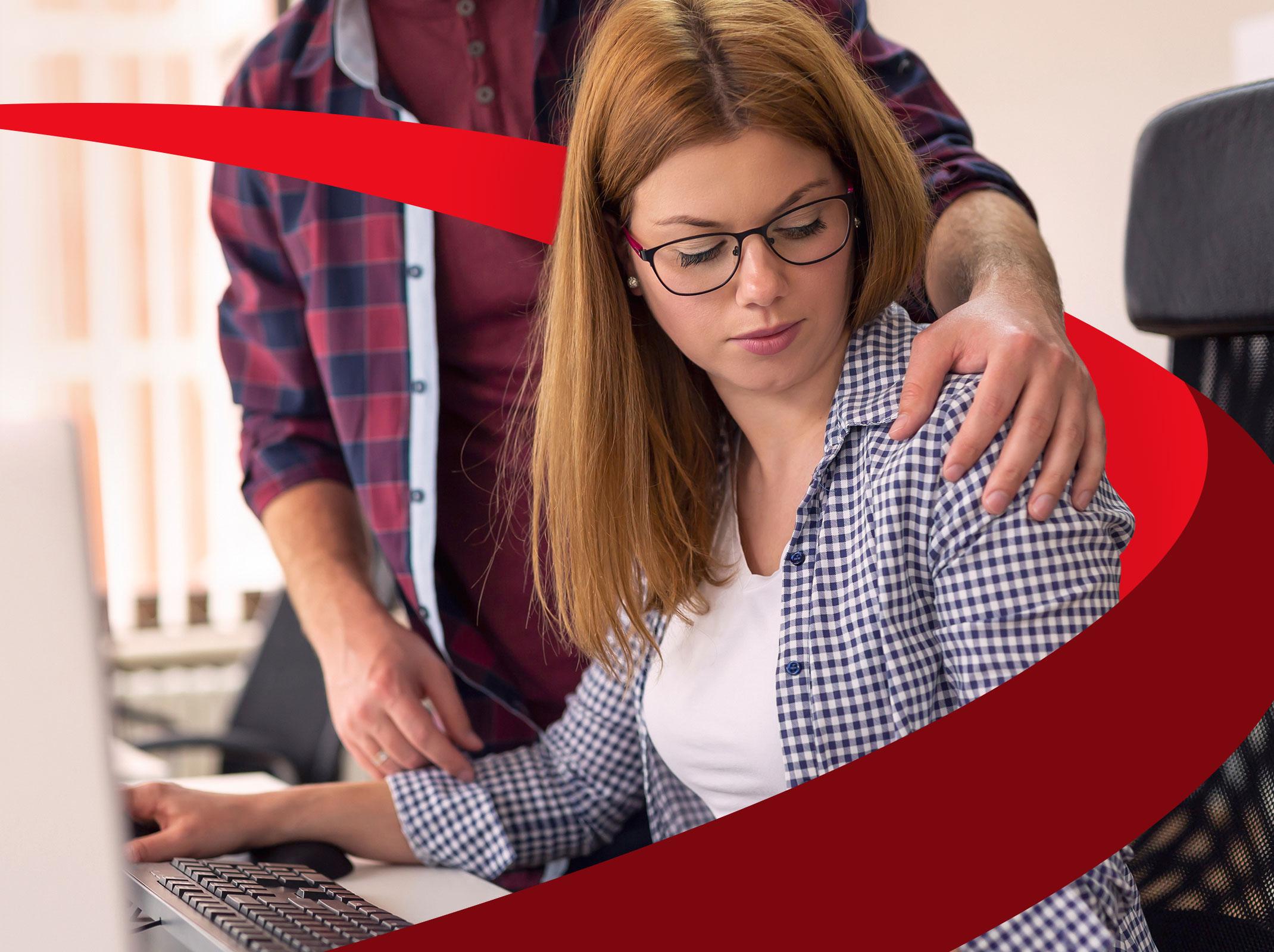 A business professional is seated at a computer. Another coworker has their hand on her shoulder and she is looking at it uncomfortably.