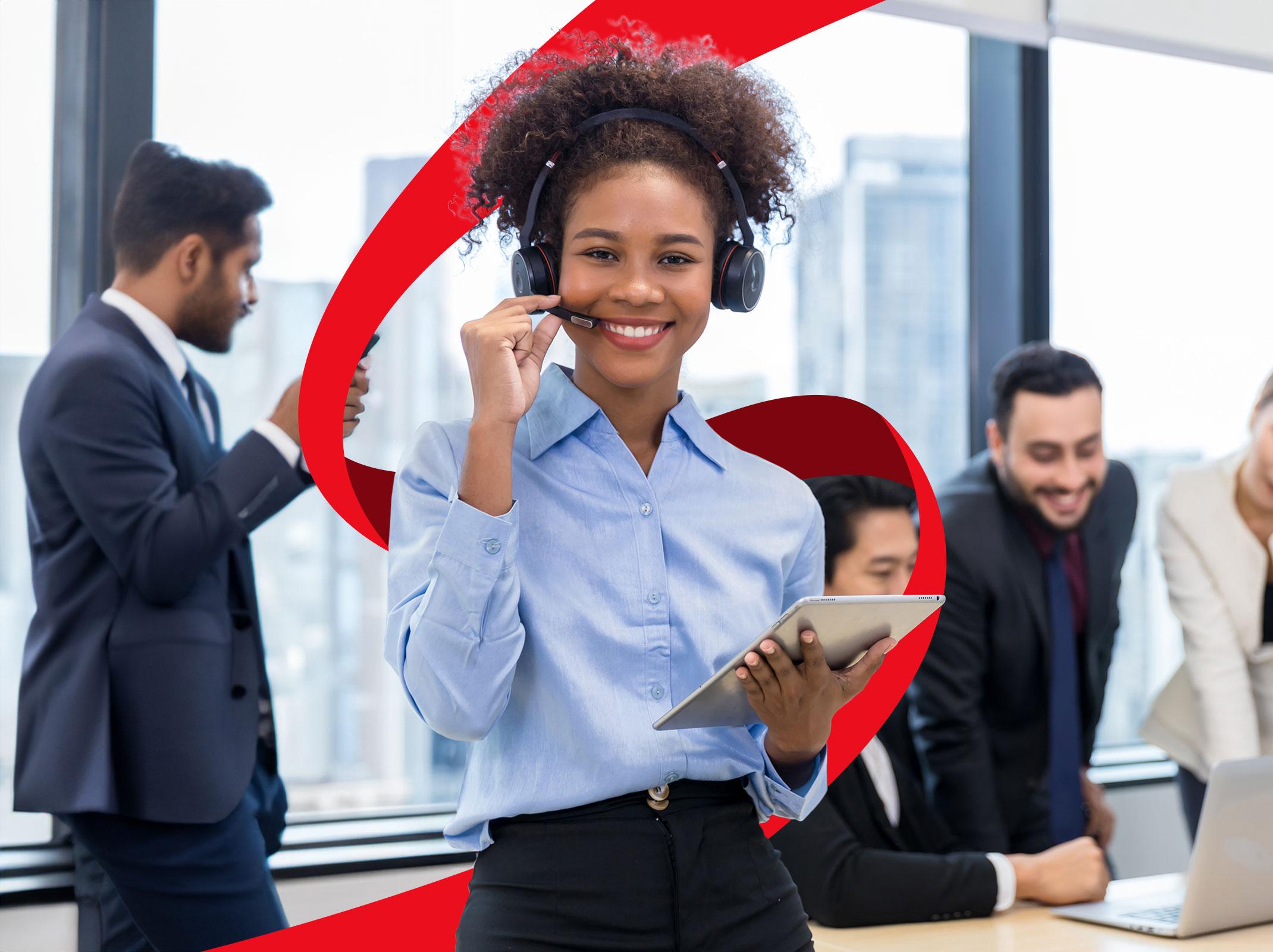 A professional sales person standing in front of a group of coworkers on a headset smiling with a tablet in her hand.