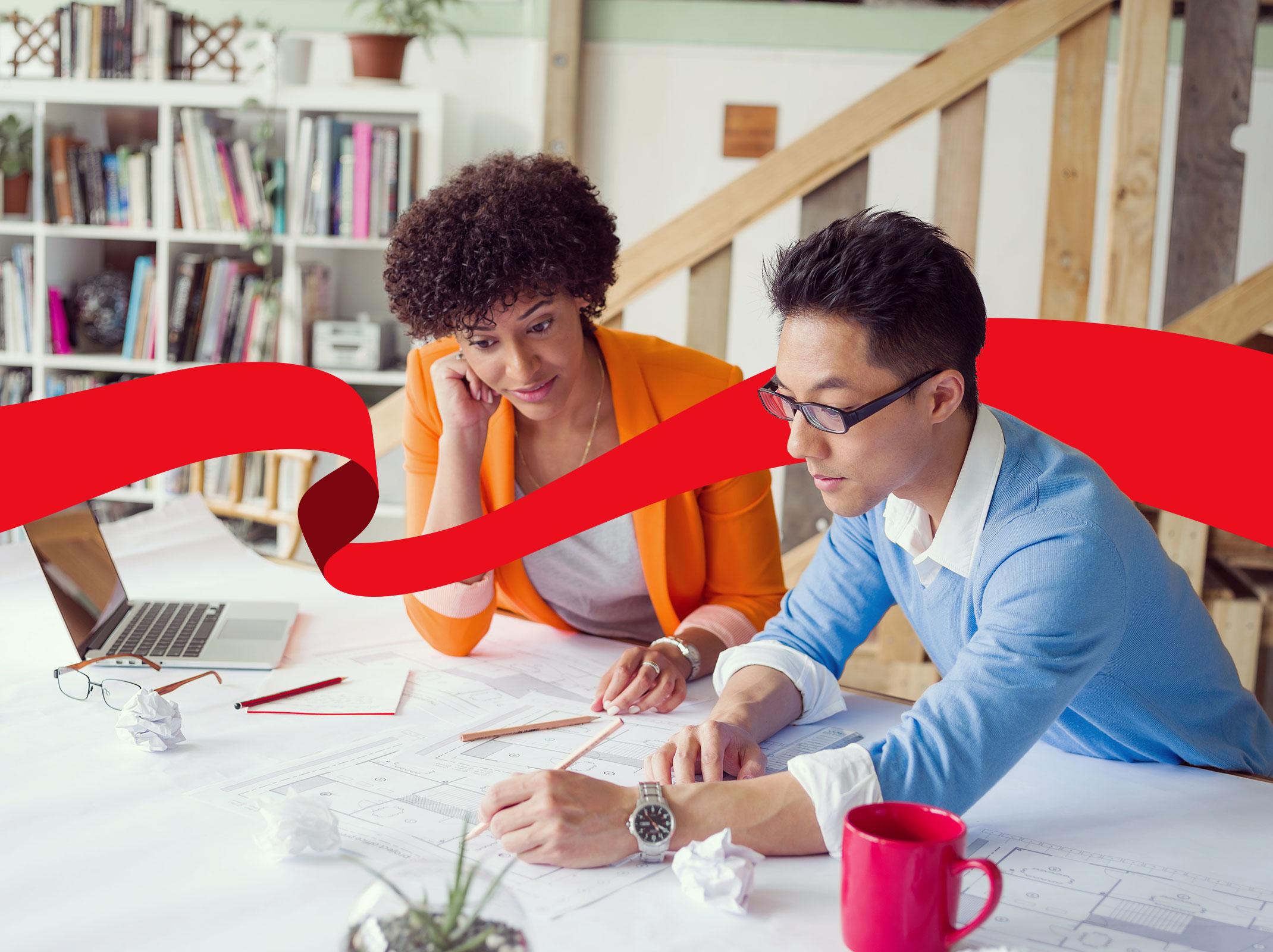 Two business professionals sitting at a table in an office setting.  They are strategically working on a document and a laptop.