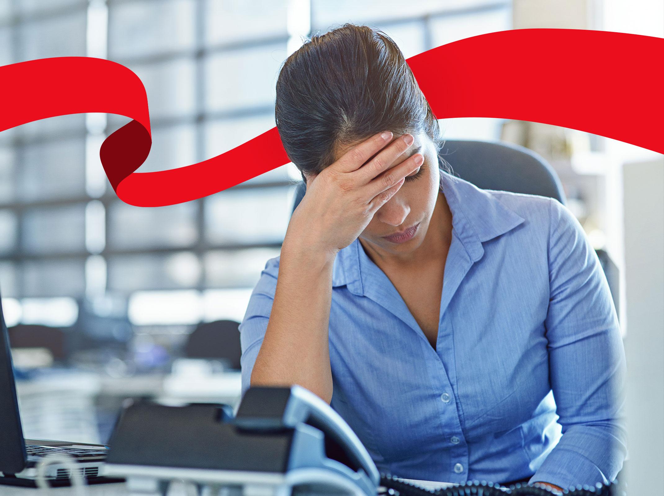A stressed professional with her hand on her forehead looking down at a desk in an office setting.
