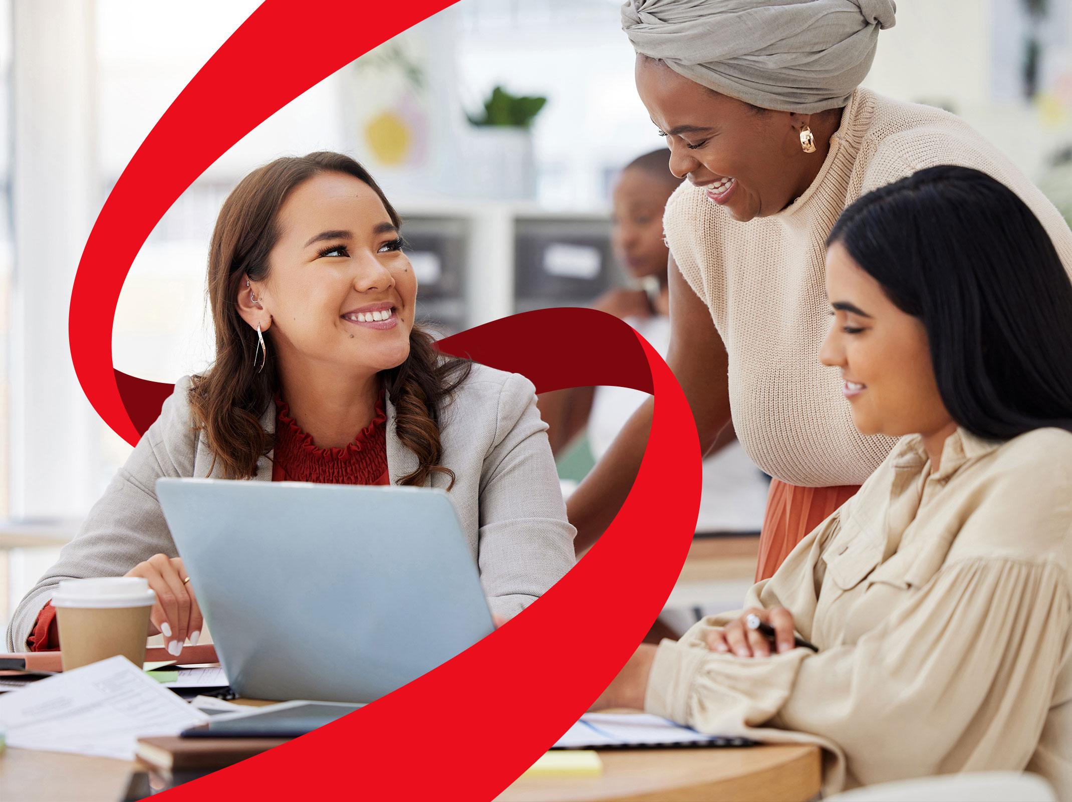 Three professional women in a business setting talking around a computer.  They are smiling.  One is standing and two are sitting.