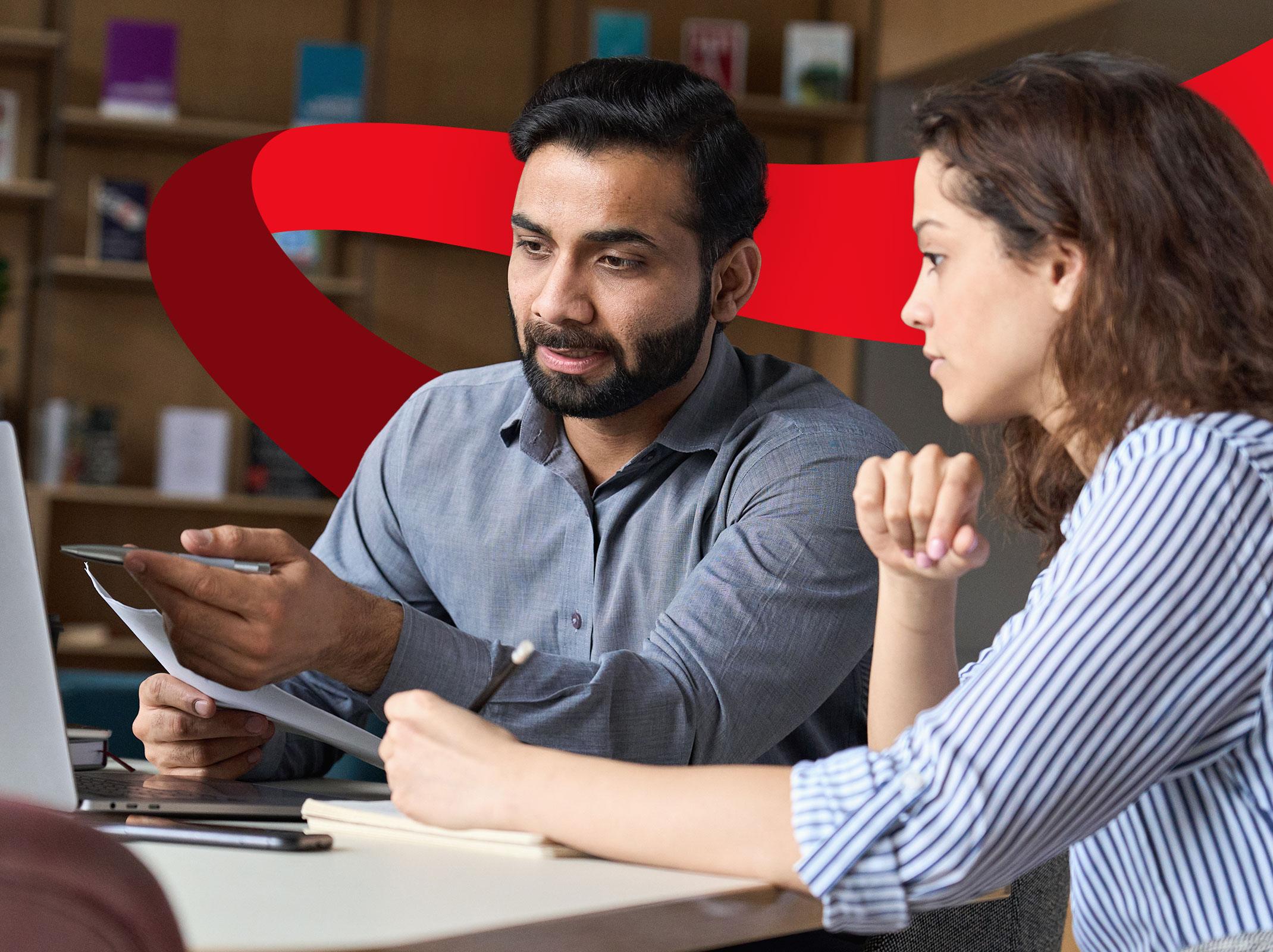 Two business professionals, one appears to be a manager, reviewing documents over a laptop in a business setting.