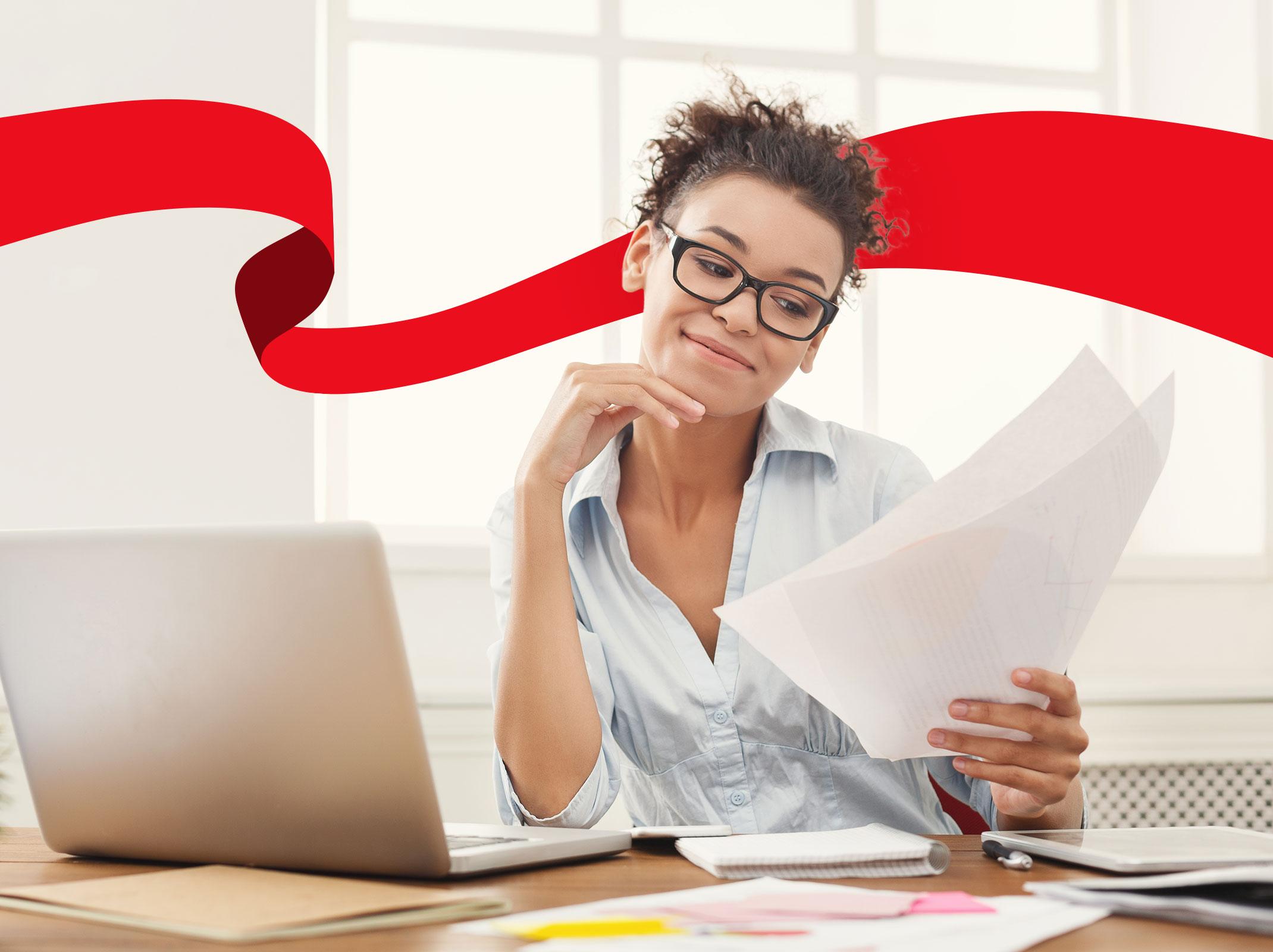 Female business professional smiling in front of a laptop.  She appears to be writing in an office setting.