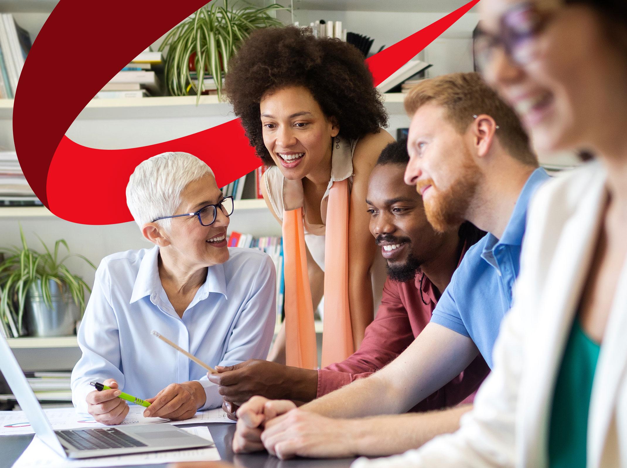 A group of business professionals that appear to be a team sitting around a conference table in a meeting room. They are engaged in a discussion, with one man speaking while others listen attentively. The table is covered with documents, laptops, and coffee cups.