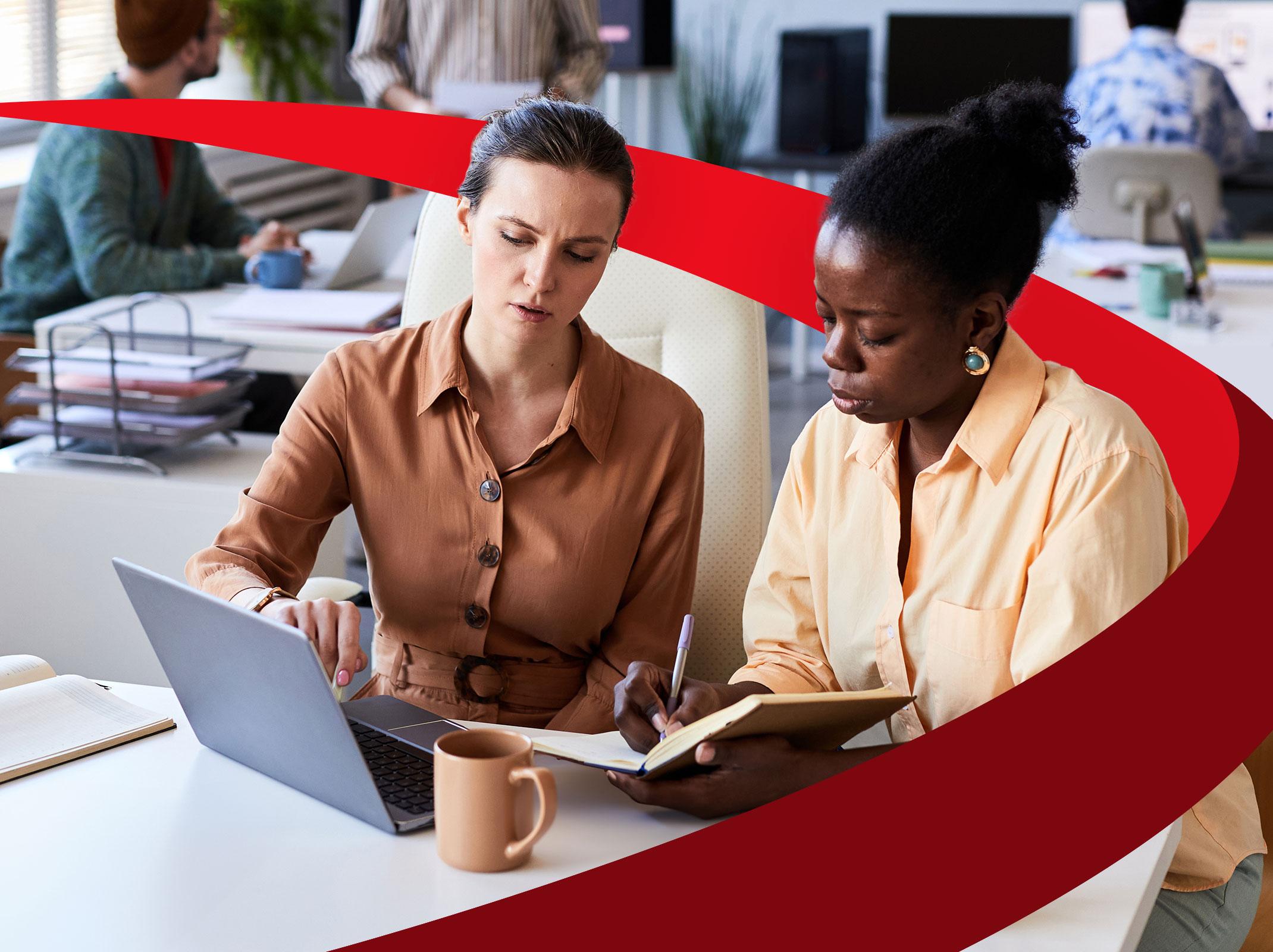 A business leaders is presenting something to another business professional on her laptop.  The second professional is taking notes in a notebook.