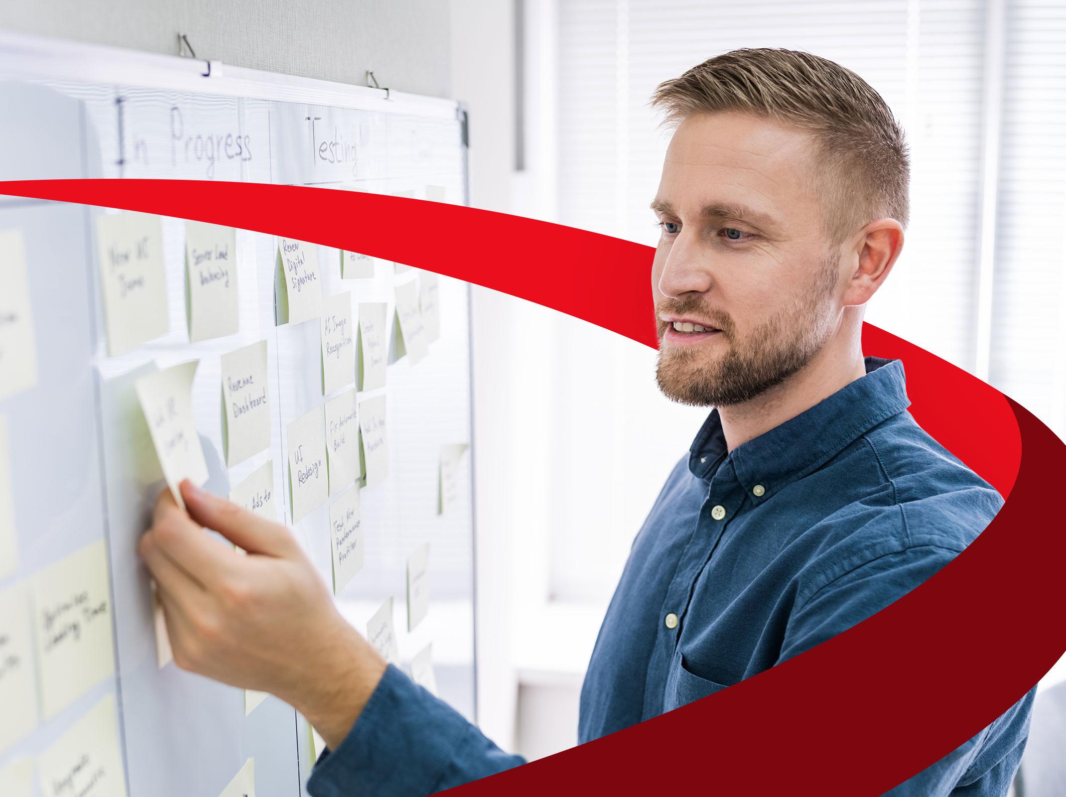 A project manager standing in front of a board of tasks.  He is moving sticky notes around on the board in a business setting.