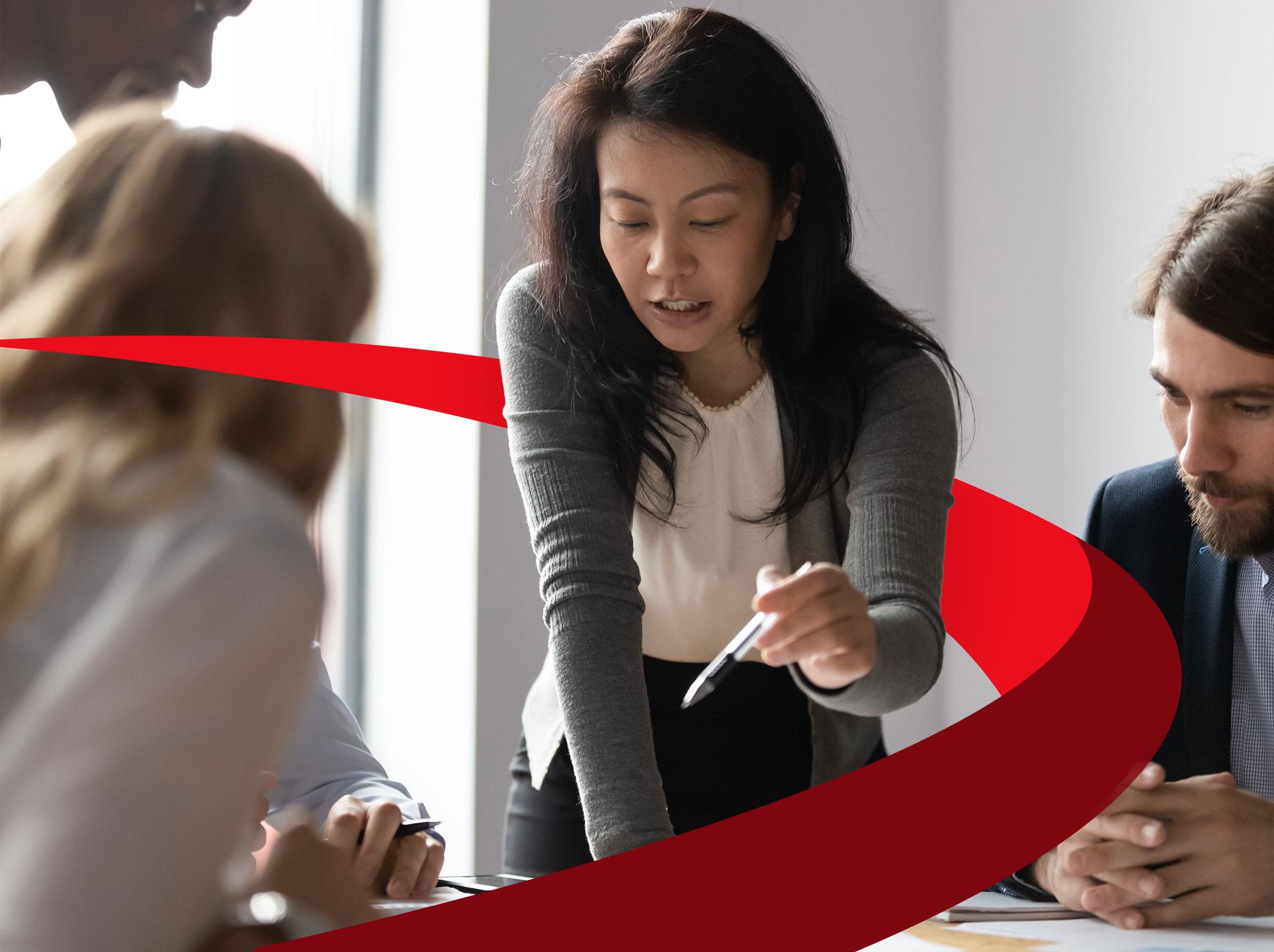 A leader directing a team of two other coworkers around a table in a business setting.  The leader is standing and pointing at something on the table.